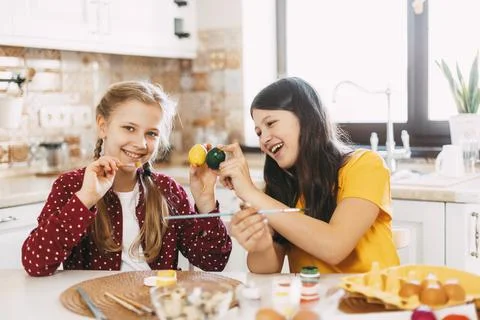 Two sisters are sitting at the table and painting Easter eggs in different Stock Photos