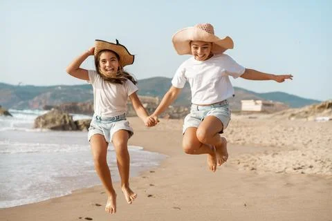 Two sisters jumping while holding hands on a sandy beach Stock Photos