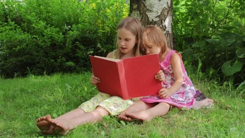 Two sisters reading a red book together in a sunny garden setting Video stock 298184749