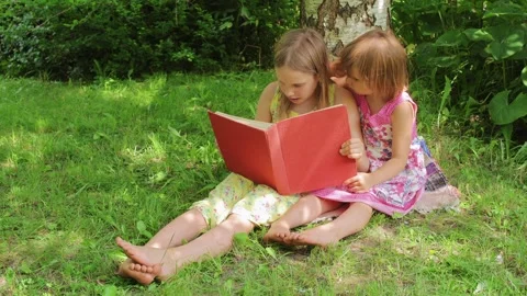 Two sisters reading a red book together in a sunny garden setting Stock Footage 298184759