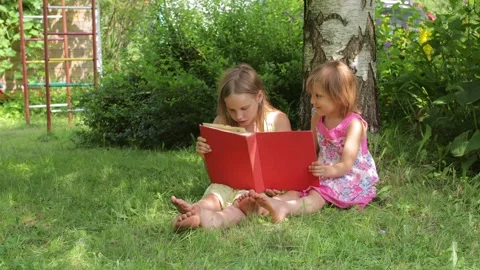 Two sisters reading a red book together in a sunny garden setting Stock Footage 298185500