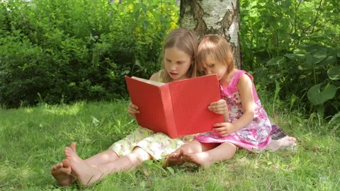 Two sisters reading a red book together in a sunny garden setting Stock Footage 298186007