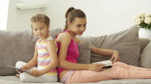 Two sisters sitting on the couch with a book and a tablet Stock Footage 24568214