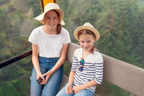 Two sisters smiling inside mountain cable car with summer hats, casual outf.. Stock Photos