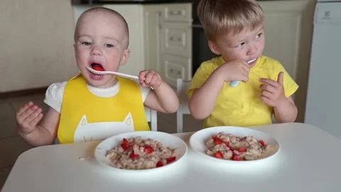 Two small children eating milk porridge sitting at a table in the kitchen at Stock Footage 243813956
