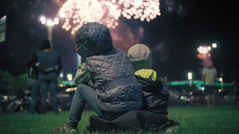Two small children sit on the grass in the evening and watch fireworks. Salut Stock Footage 99577076