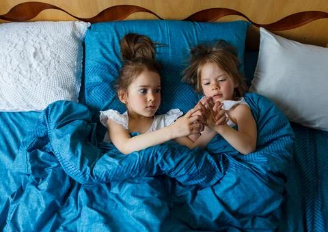 Two small smiling cute sisters are lying in bed, top view. Stock Photos