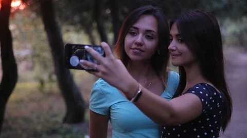 Two smiley cheerful girlfriends doing photos in the evening park Video stock 100750090