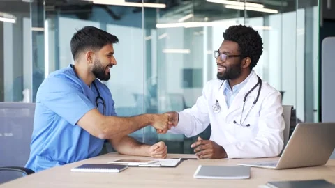 Two smiling doctors sitting at desk in hospital office talking and shaking hands Stock Footage 329052478