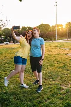 Two smiling friends taking a selfie, in a public park in the city at sunset Stock Photos