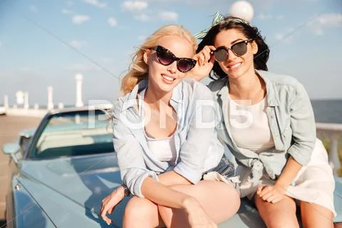 Two smiling women sitting on the car hood in summer Stock Photos