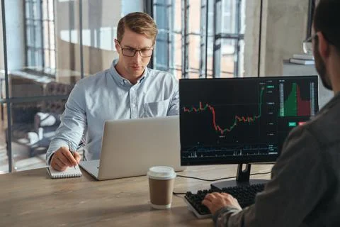 Two stock traders co-working at office sitting at table in front of computers Stock Photos
