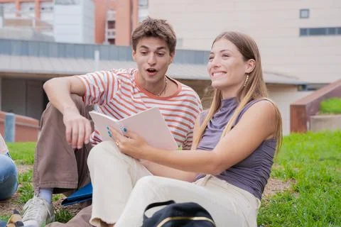 Two students on campus sitting surprised reviewing notes for exam outdoors. Stock Photos