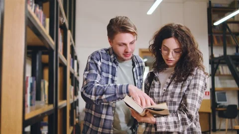 Two students discussing a book in a library with shelves filled with literature Stock Footage 318094290