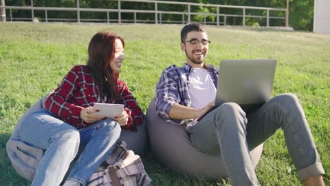 Two students sitting on pillows in the grass, an Asian female in a red plaid Stock Footage 283841165