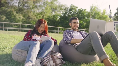 Two students sitting on pillows in the grass, an Asian female in a red plaid Stock Footage 283841200