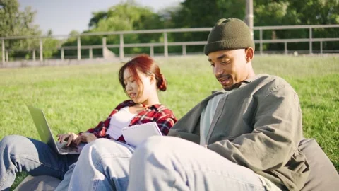 Two students sitting on pillows in the grass, an Asian female in a red plaid Stock Footage 283841403