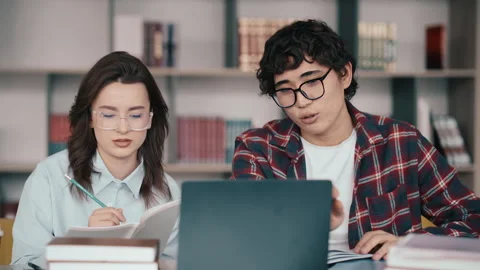Two students studying together at a library desk with laptop and books Stock Footage 341889307