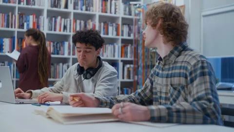 Two Students Talking and Working on Homework as Another Browses a Bookshelf 库存影片 335125290