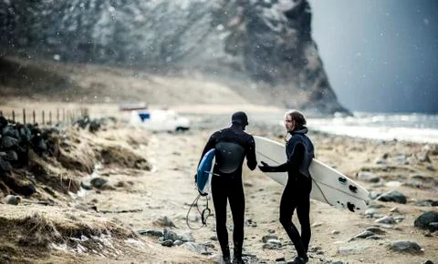 Two surfers wearing wetsuits and carrying surfboards walking towards a van. Stock Photos