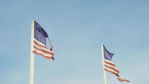 Two tattered American flags waving in the wind against blue sky. Low angle view. Stock Footage 321344592