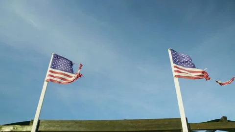 Two tattered American flags waving in the wind against blue sky. Low angle view. Stock Footage 321344901