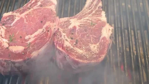 Two thick juicy raw ribeye steaks grilling on a charcoal barbecue with smoke ris Stock Footage 338213091