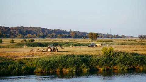 Two tractors make hay in a spacious meadow by the river, with hills and for.. Stock Photos