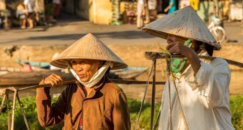 Two Vietnamese women with conical hats, carrying shoulder poles by a river in Stock Photos