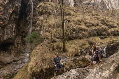 Two Viking explorers rest on the rocks next to a waterfall in the forest Stock Photos
