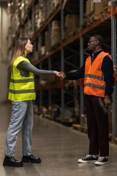 Two warehouse workers wearing safety vests shake hands in a busy distribution Stock Photos