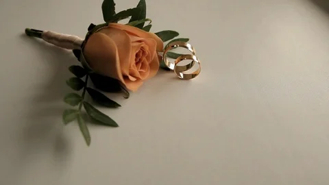 Two wedding rings roll on a white surface next to a rose Stock Footage 86987173