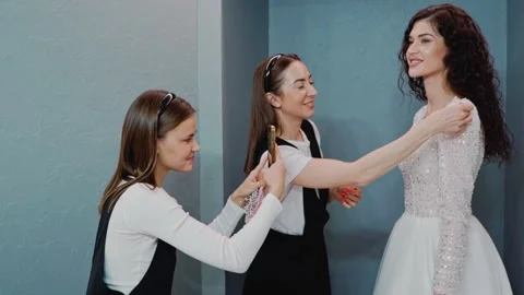 Two wedding salon consultants help the bride try on her wedding dress. One Vidéo 199851950