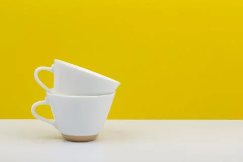 Two white coffee cups on in another on white table against yellow background Stock Photos