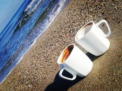Two white cups of coffee stand on gray sand near the sea Stock Photos