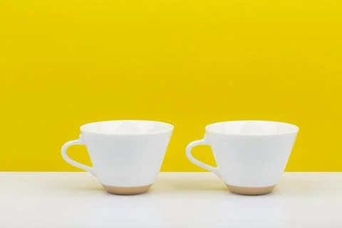 Two white glossy ceramic cups on white table against bright yellow background Stock Photos