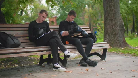 Two white men on bench reviewing route notes and helmet, relaxed autumn park Video stock 325032136