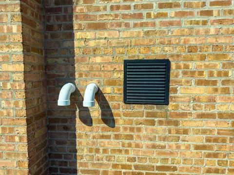 Two white pipes extend from a brick wall beside a black vent grille. The brig 스톡 사진