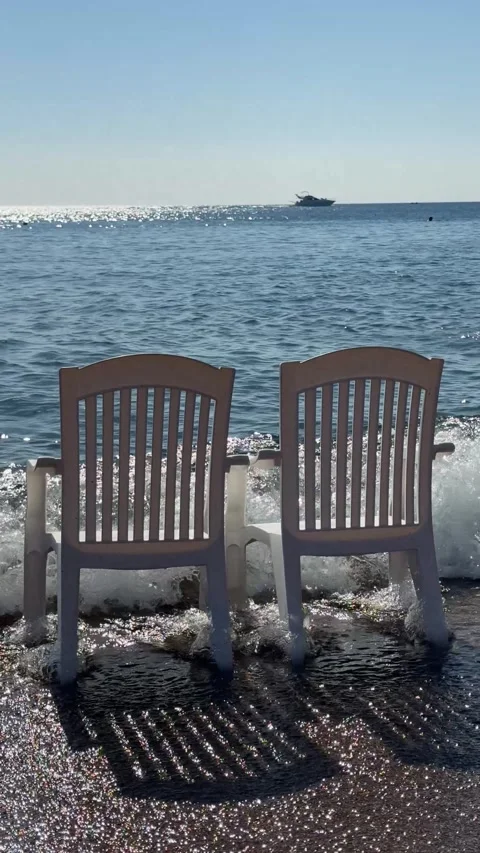 Two white plastic chairs stand in the water on a sandy beach, gently touched  Stock Footage 303359195