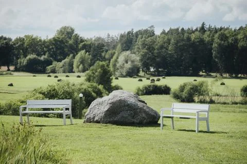 Two white wooden benches positioned near a large rock in a green field with.. Stock Photos