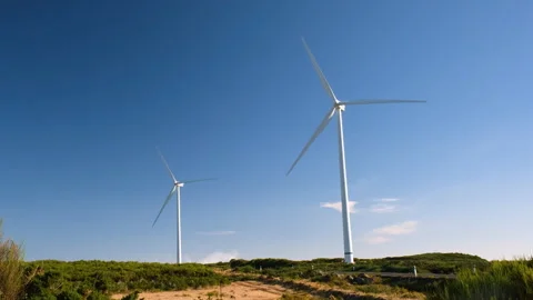 Two wind turbines generating renewable energy on a green hillside under a clear Stock Footage 346150481