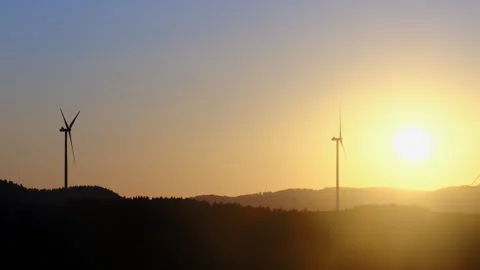 Two wind turbines rise over valley as closer turbine turns Stock Footage 323887428
