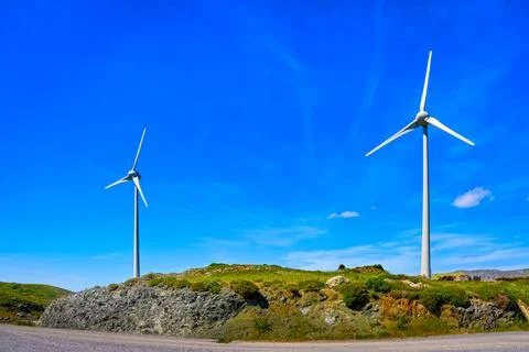 Two wind turbines by road on top of green hill on sunny day, Greece Stock Photos