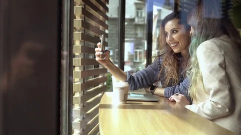 Two woman friends pose for fun selfies together in cafe. View through the window Stock Footage 78377300