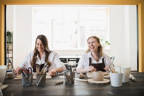 Two women in aprons sitting by table, kneading clay and making earthenware at Foto stock