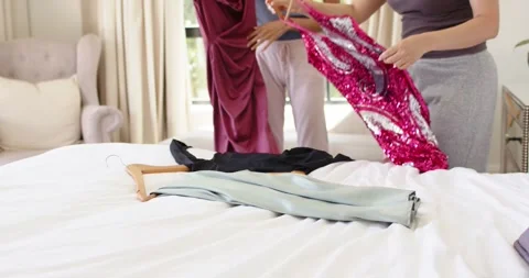 Two women are comparing maroon and sequined dresses on bed, swapping hangers to Stock Footage 335927970