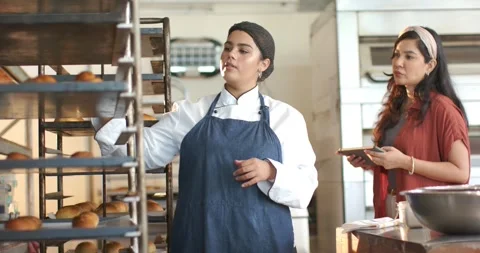 Two women at bakery rack, baker pointing, removing roll, checking quality as Stock Footage 340393857