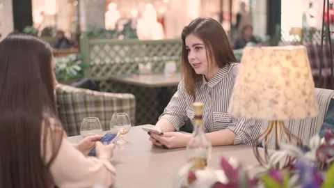 Two women in cafe using apps on phone. on the table bottle and two glasses. 库存影片 144598933