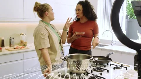 Two women co-hosts gesturing and explaining, one pouring oil into pan while Stockbeeldmateriaal 332428802