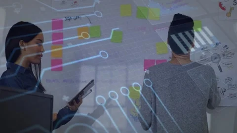 Two women at design board, left woman tapping tablet activating HUD overlay Stock Footage 330227432